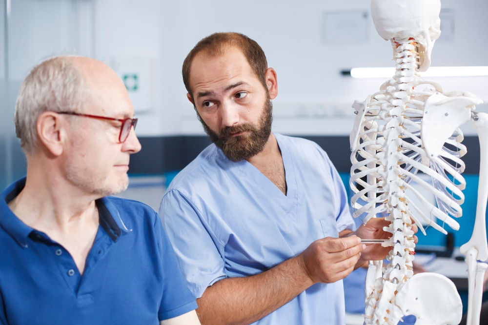 A doctor pointing to a plastic skeleton’s spine while explaining the most common causes of spinal cord injuries to a patient.